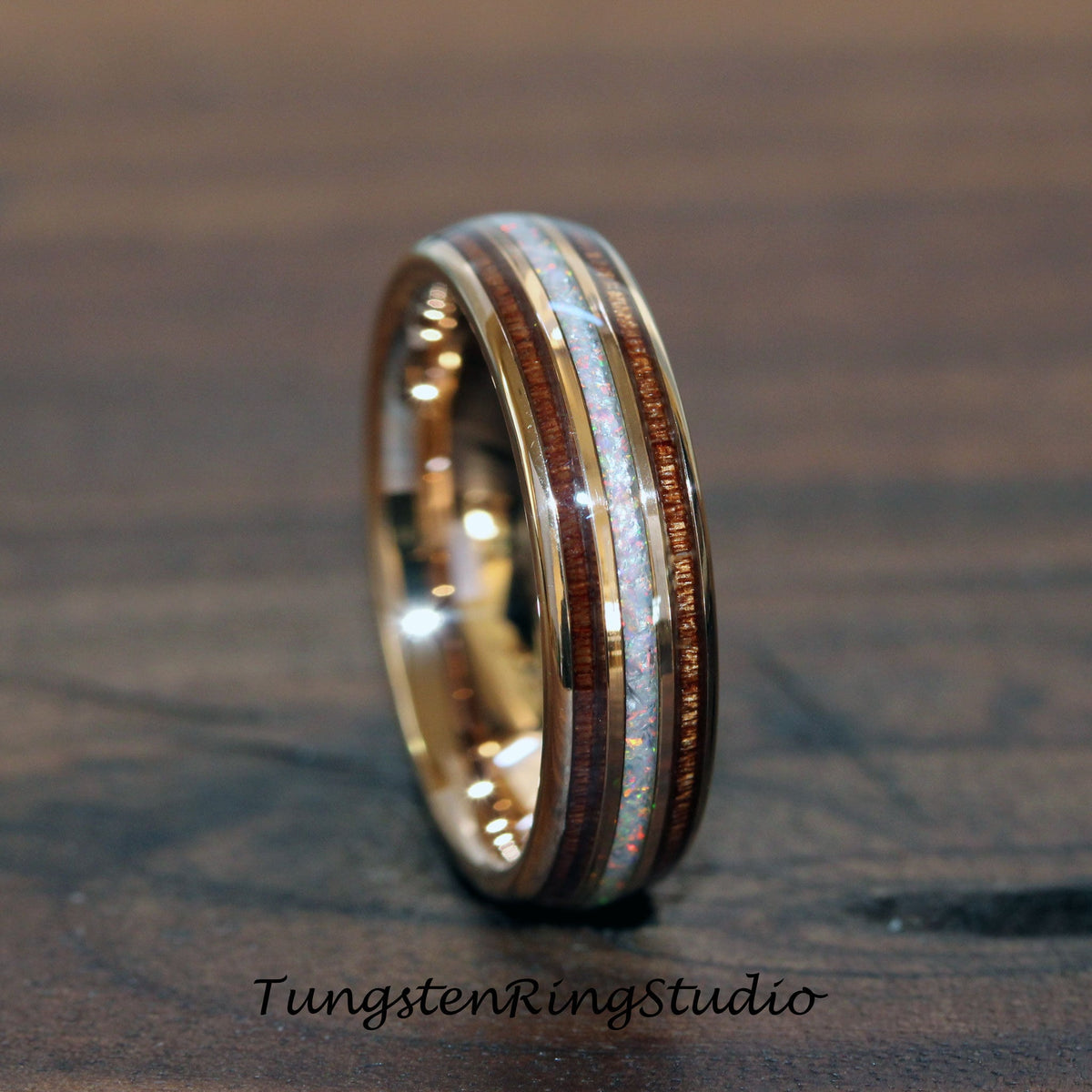 May include: A gold-colored ring with a central band of white opal, flanked by bands of dark wood. The ring has a polished finish and the text "TungstenRingStudio" is visible at the bottom.