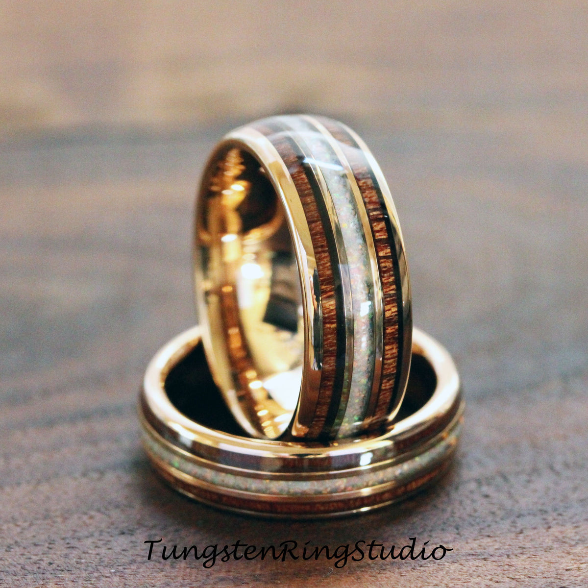 May include: Two gold-colored rings with a wood and opal inlay. The rings are stacked on top of each other, with the top ring in focus. The rings have a polished finish and are set against a dark wood background. The text "TungstenRingStudio" is at the bottom.