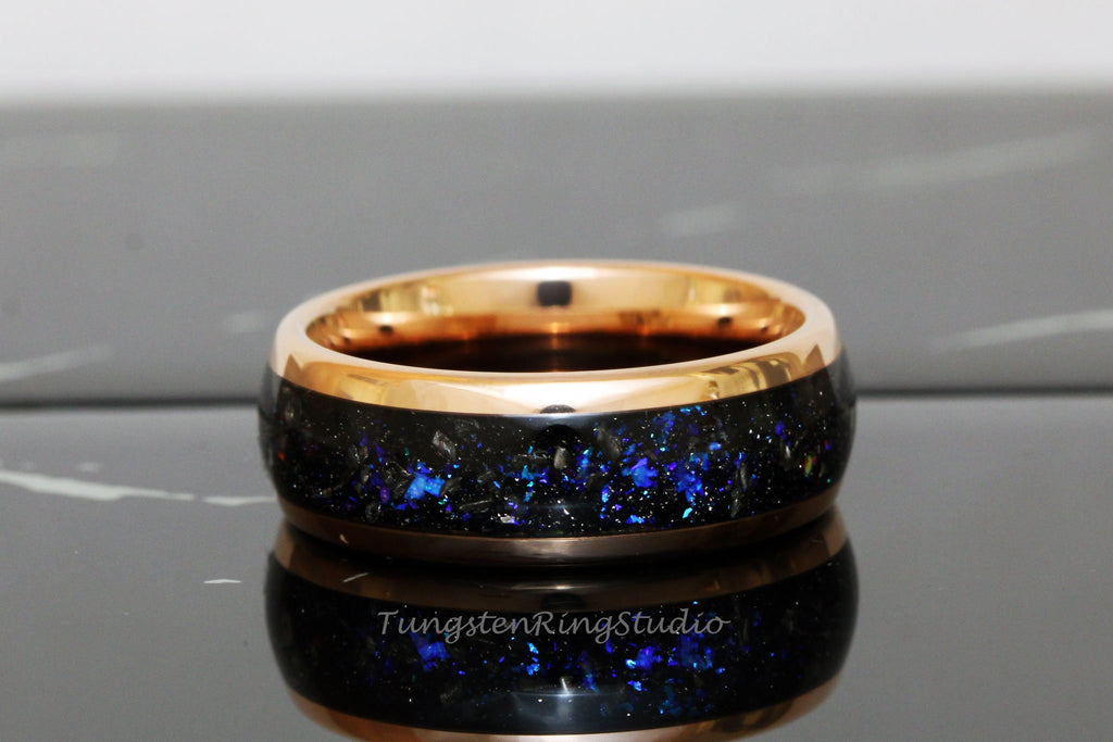 May include: A black tungsten ring with a rose gold inlay and blue glitter embedded in the black band. The ring is on a black surface.