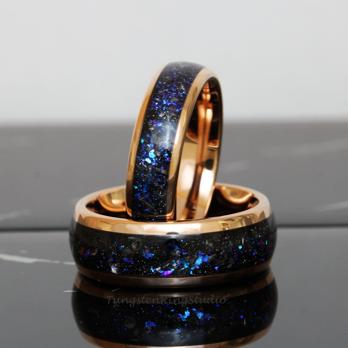 May include: Two tungsten carbide rings with a black and blue inlay and a rose gold band. The rings are stacked on top of each other.