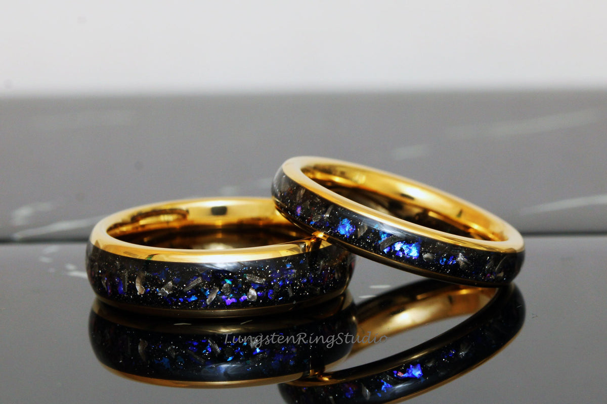 May include: Two gold tungsten carbide rings with a black inlay and blue and silver glitter. The rings are stacked on top of each other.
