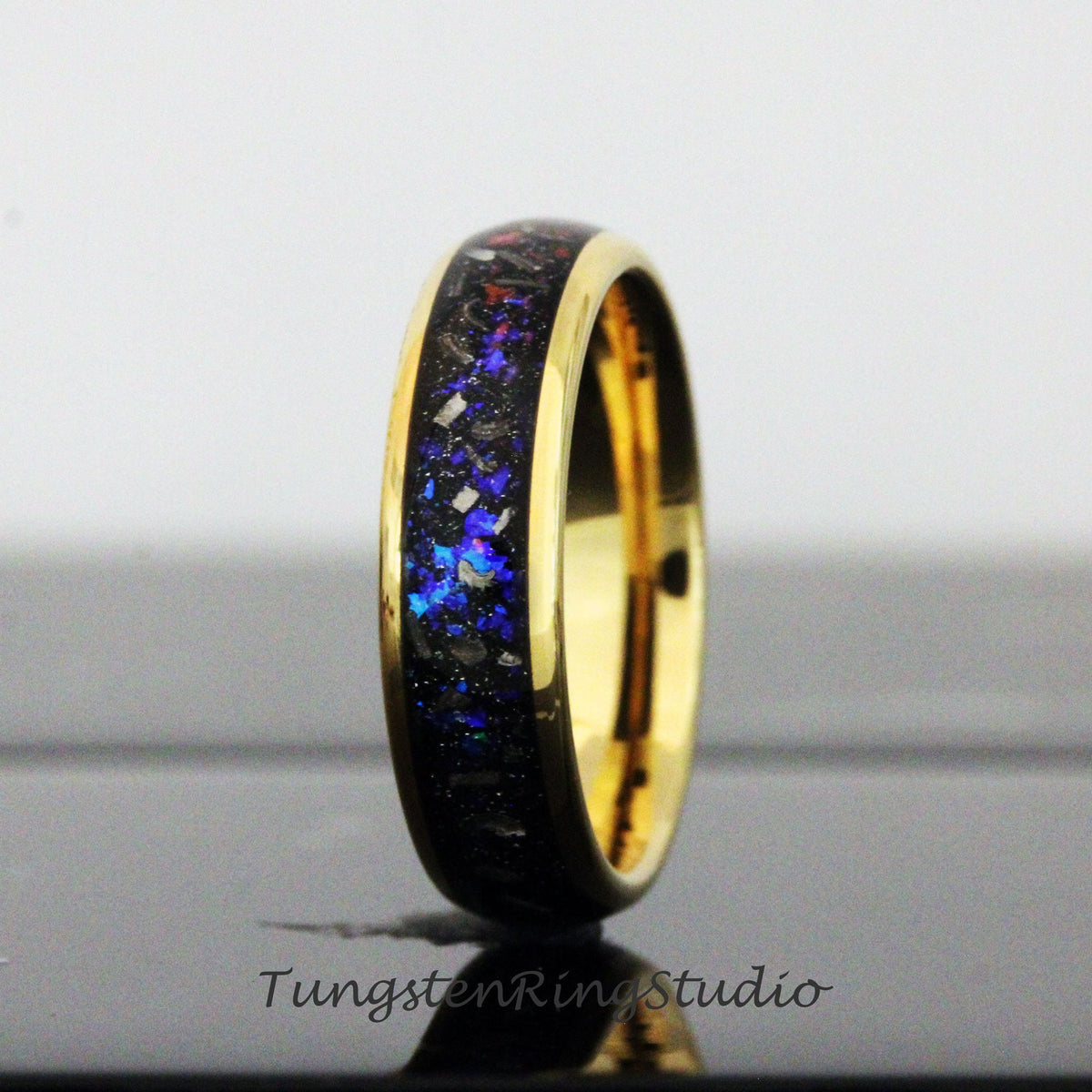 May include: A gold tungsten carbide ring with a black and blue inlay. The inlay has a speckled pattern of blue, black, and silver.