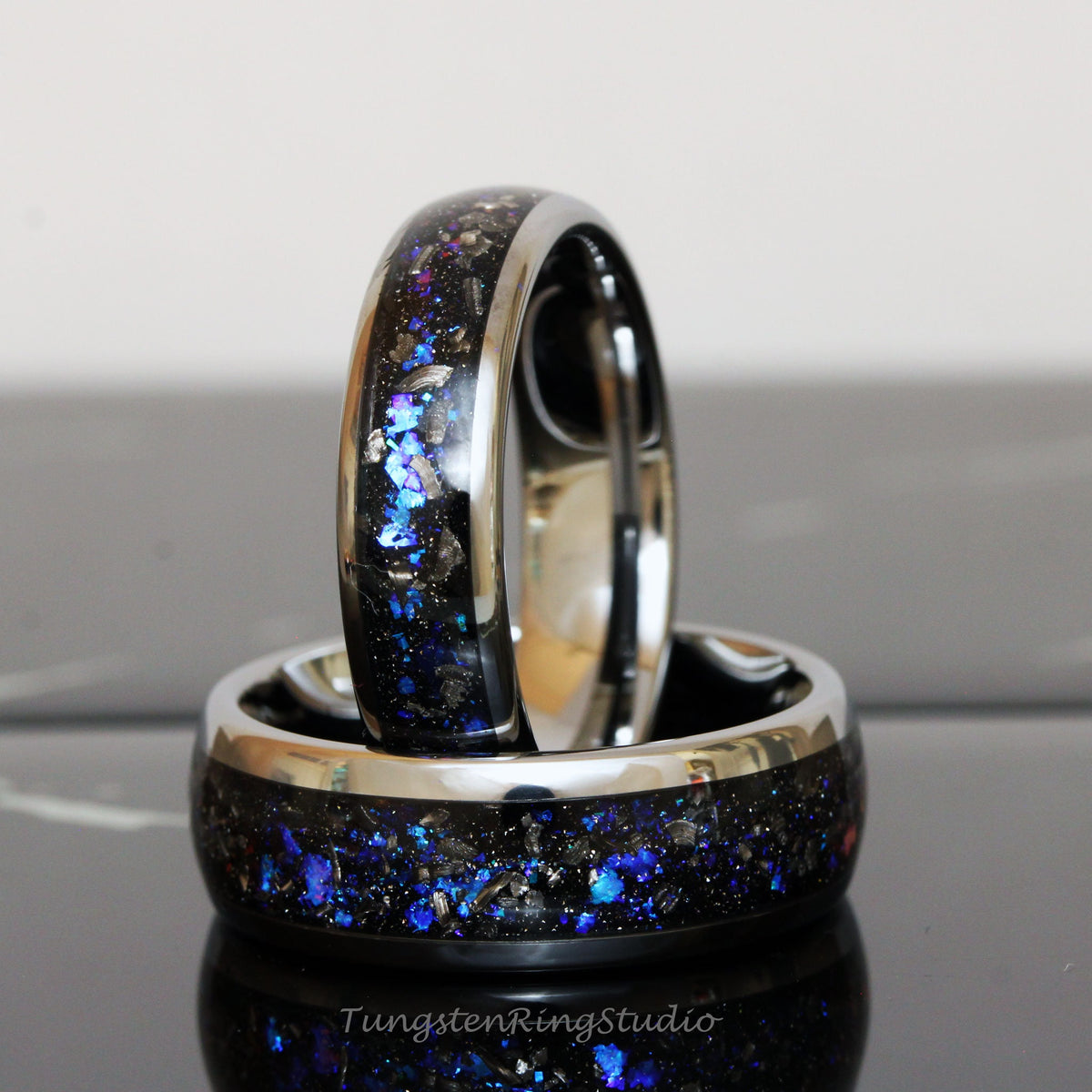 May include: Two black tungsten carbide rings with a silver band and blue and silver glitter embedded in the black surface. The rings are stacked on top of each other.