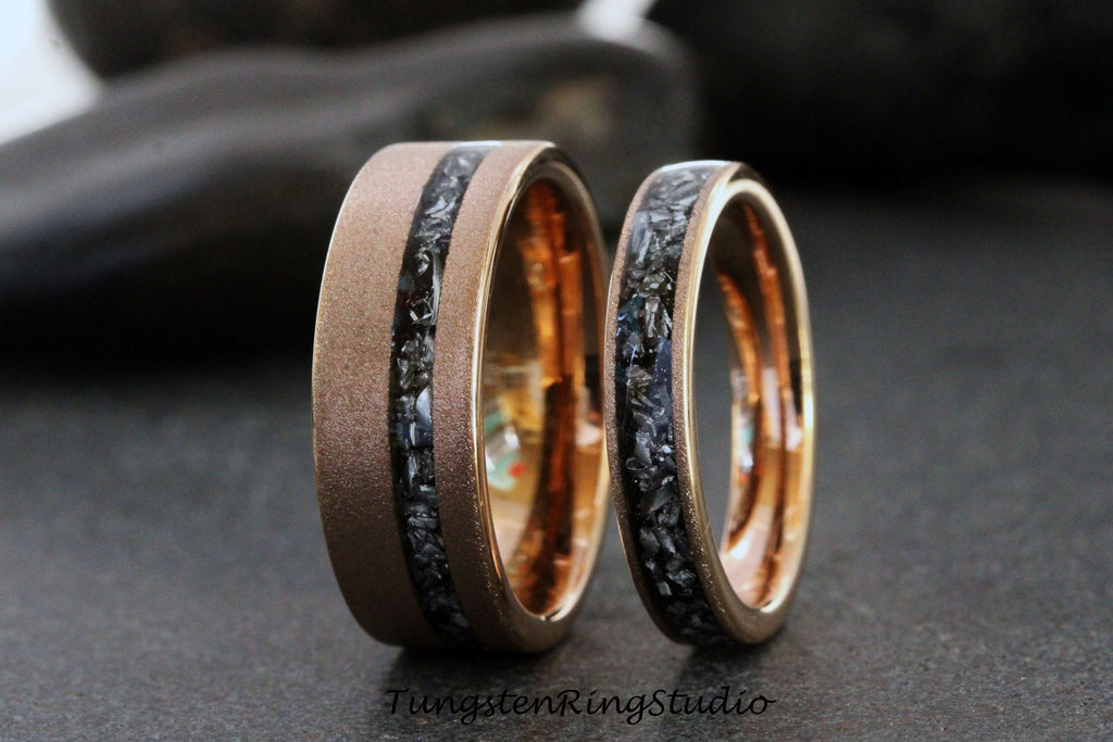 May include: Two wedding rings with a brushed bronze finish and a black stone inlay. The rings have a polished gold interior. The larger ring is wider than the smaller ring. The rings are displayed on a dark surface.