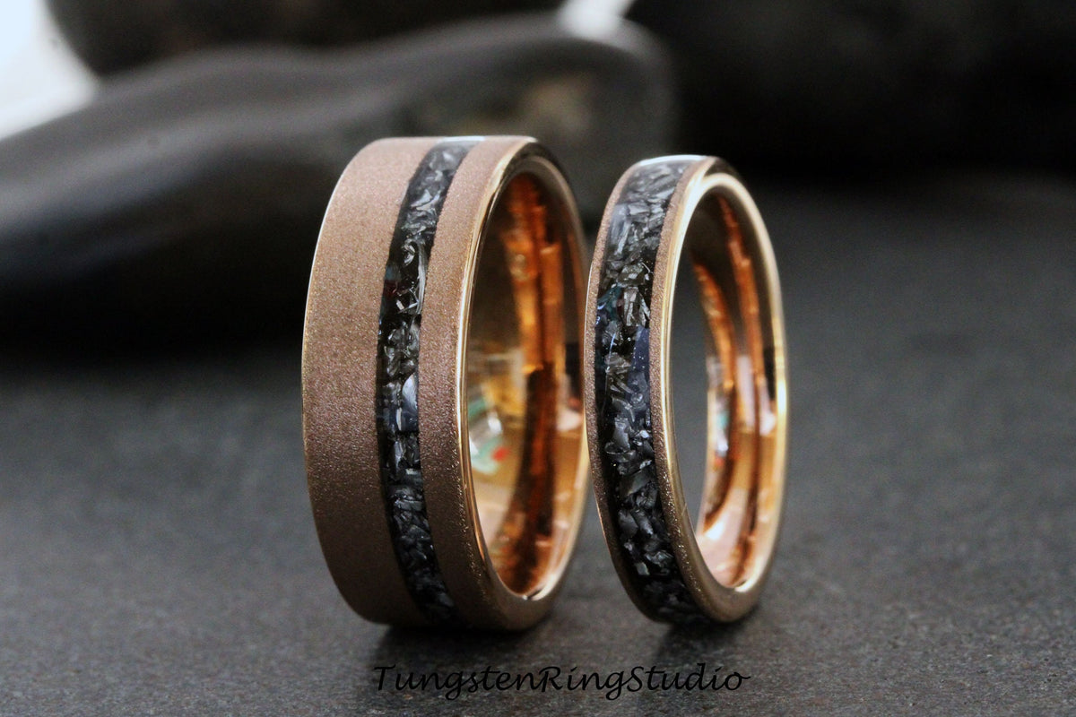 May include: Two wedding rings with a brushed bronze finish and a black stone inlay. The rings have a polished gold interior. The larger ring is wider than the smaller ring. The rings are displayed on a dark surface.