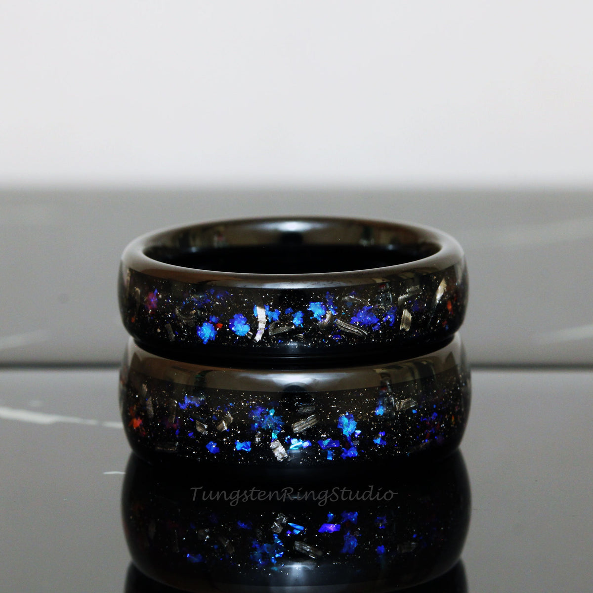 May include: Two black tungsten carbide rings with blue and silver glitter embedded in the black resin. The rings are stacked on top of each other.