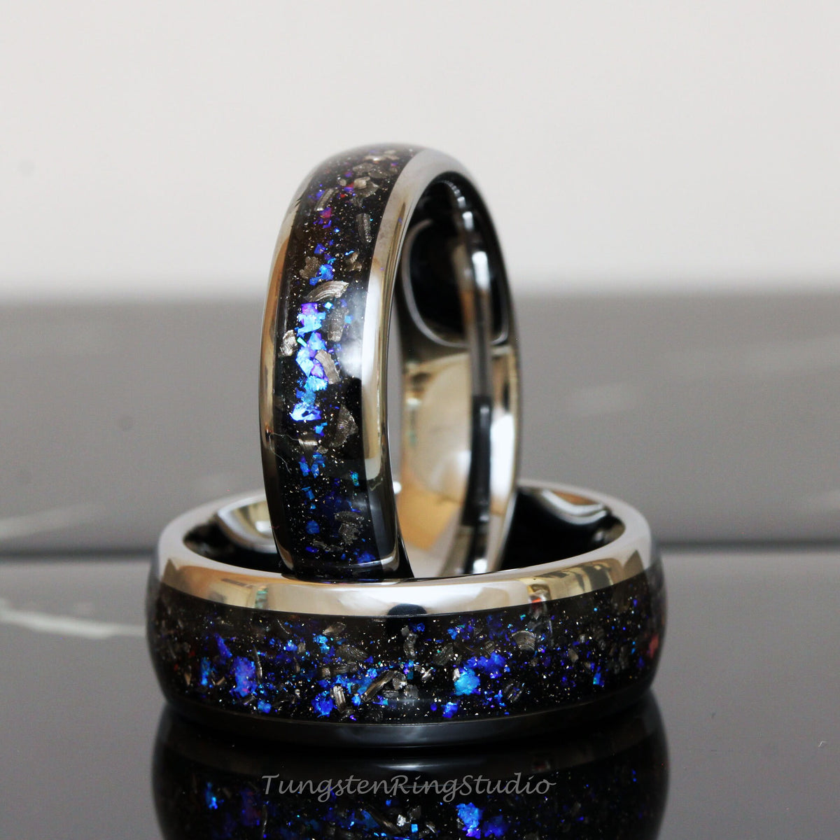 May include: Two black tungsten carbide rings with a silver band and blue and silver glitter embedded in the black surface. The rings are stacked on top of each other.