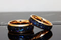 May include: Two matching tungsten carbide rings with a black inlay and a rose gold band. The inlay has a sparkly blue and black pattern.