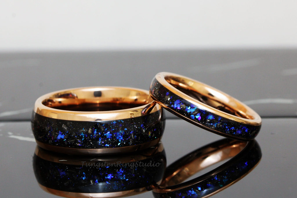 May include: Two matching wedding bands with a rose gold finish. The bands are made of tungsten carbide and feature a black inlay with blue glitter.