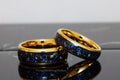 May include: Two gold tungsten carbide rings with a blue and black inlay. The rings have a gold band around the edge.