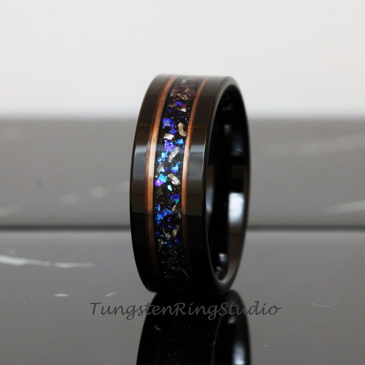 May include: A black tungsten carbide ring with a copper inlay and a blue and silver glitter inlay. The ring is on a black surface.