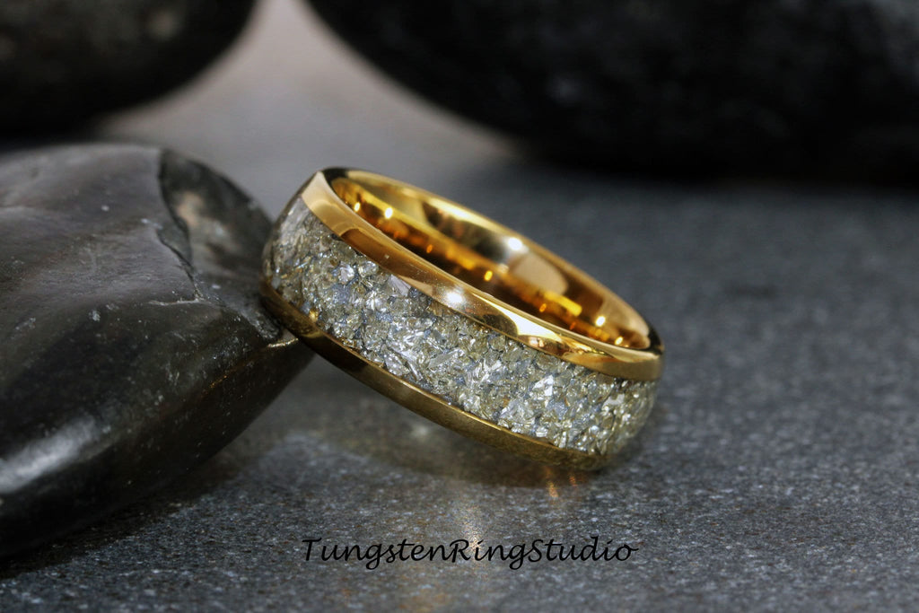 May include: A gold tungsten carbide ring with a white inlay of crushed meteorite. The ring is on a gray surface.