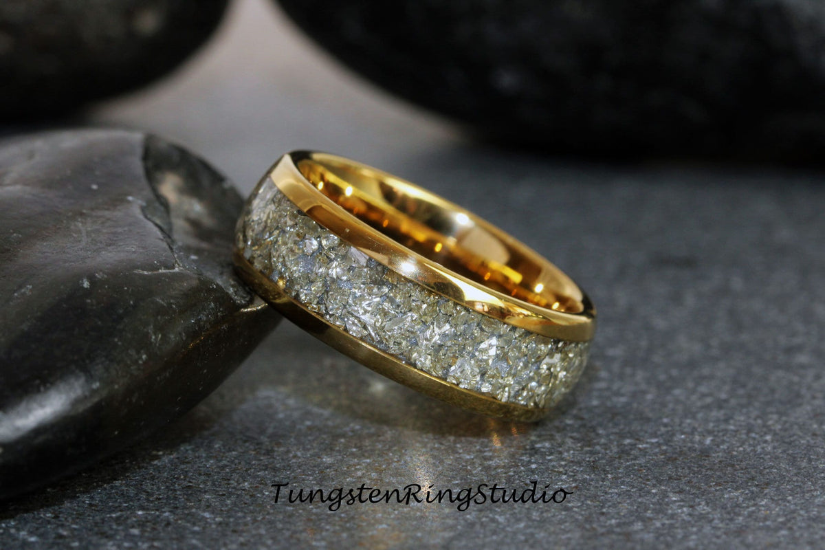 May include: A gold tungsten carbide ring with a white inlay of crushed meteorite. The ring is on a gray surface.
