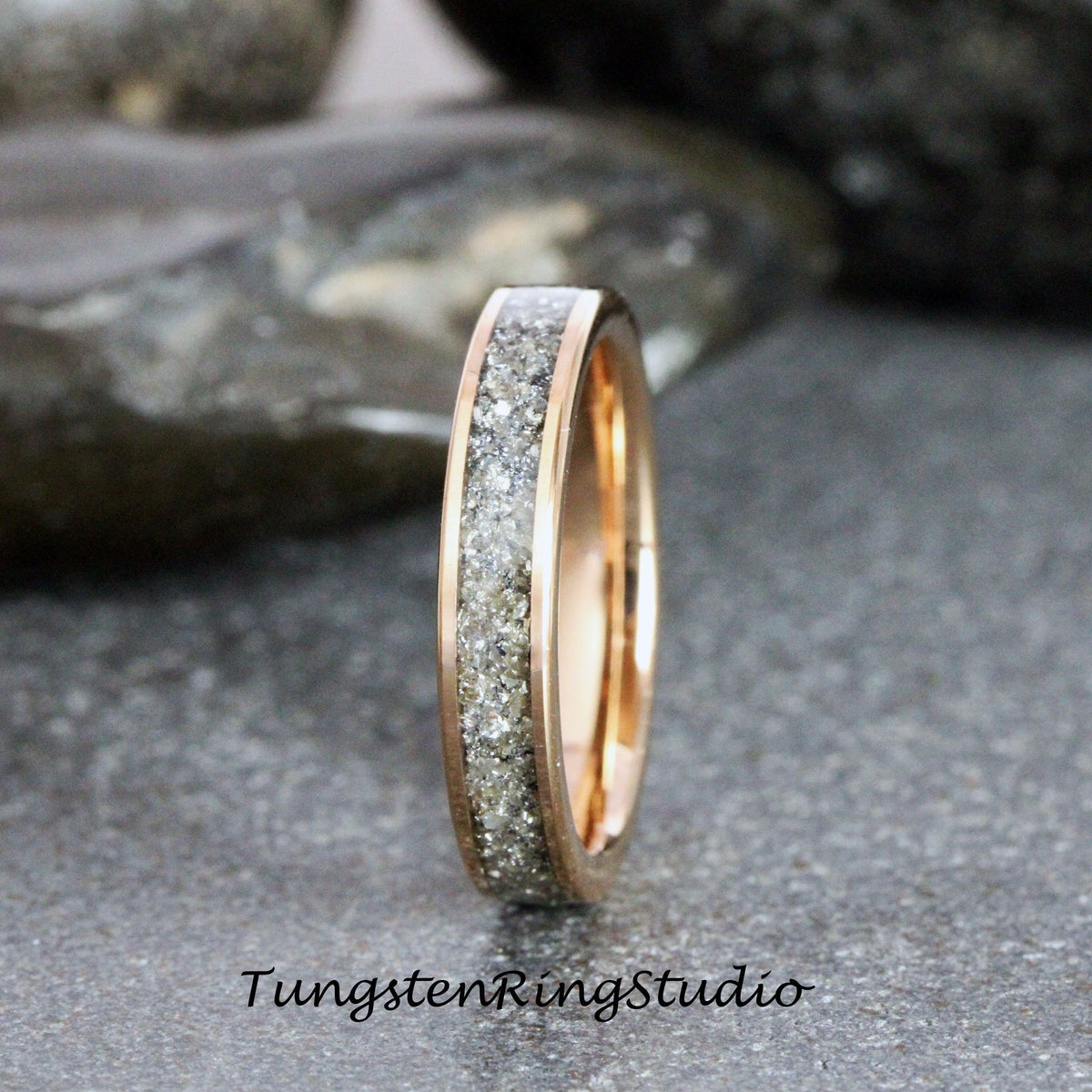 May include: A rose gold tungsten carbide ring with a band of embedded gray diamonds. Tungsten Ring Studio is printed below the ring.