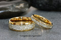 May include: Two gold tungsten carbide wedding bands with a diamond-like inlay. The bands are on a gray surface.