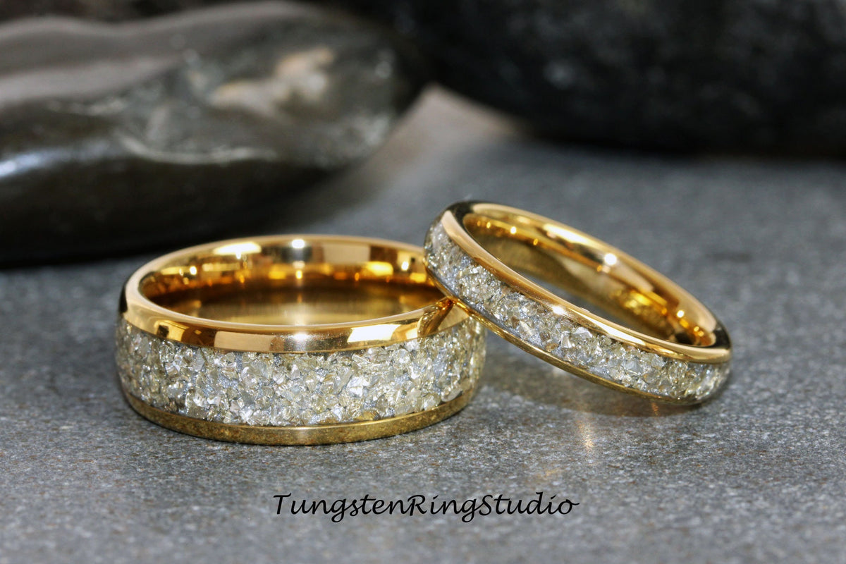 May include: Two gold tungsten carbide wedding bands with a diamond-like inlay. The bands are on a gray surface.