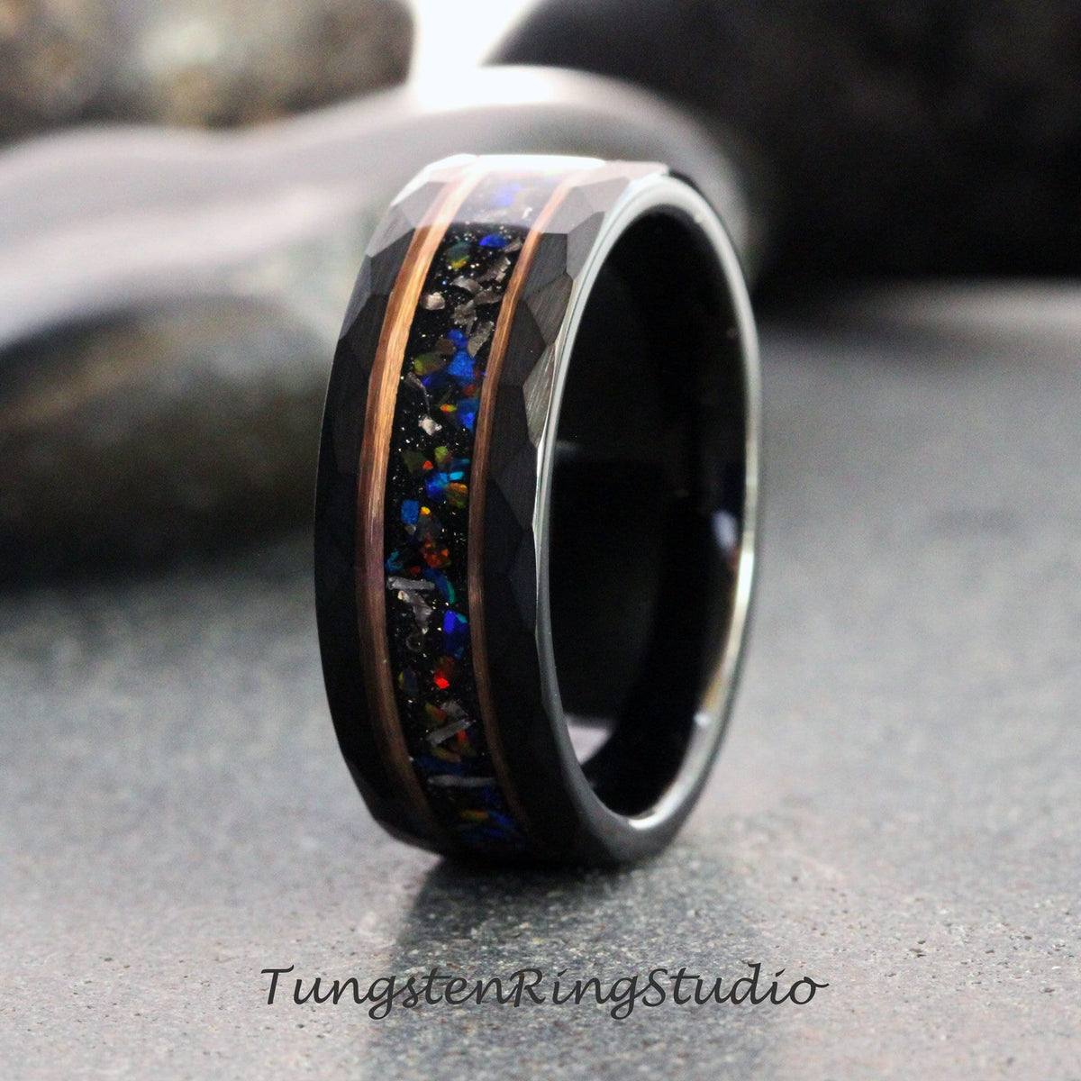 May include: A black tungsten ring with a faceted exterior and a central inlay of colorful, sparkling material. The inlay is framed by copper-colored metal bands. The ring is displayed on a gray surface. The text "TungstenRingStudio" is at the bottom.