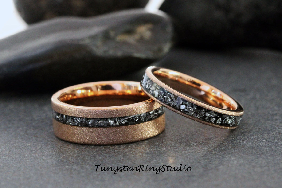 May include: Two rose gold wedding rings. The wider ring has a textured finish and a band of dark gray stones. The thinner ring has a similar design. The text "TungstenRingStudio" is visible.