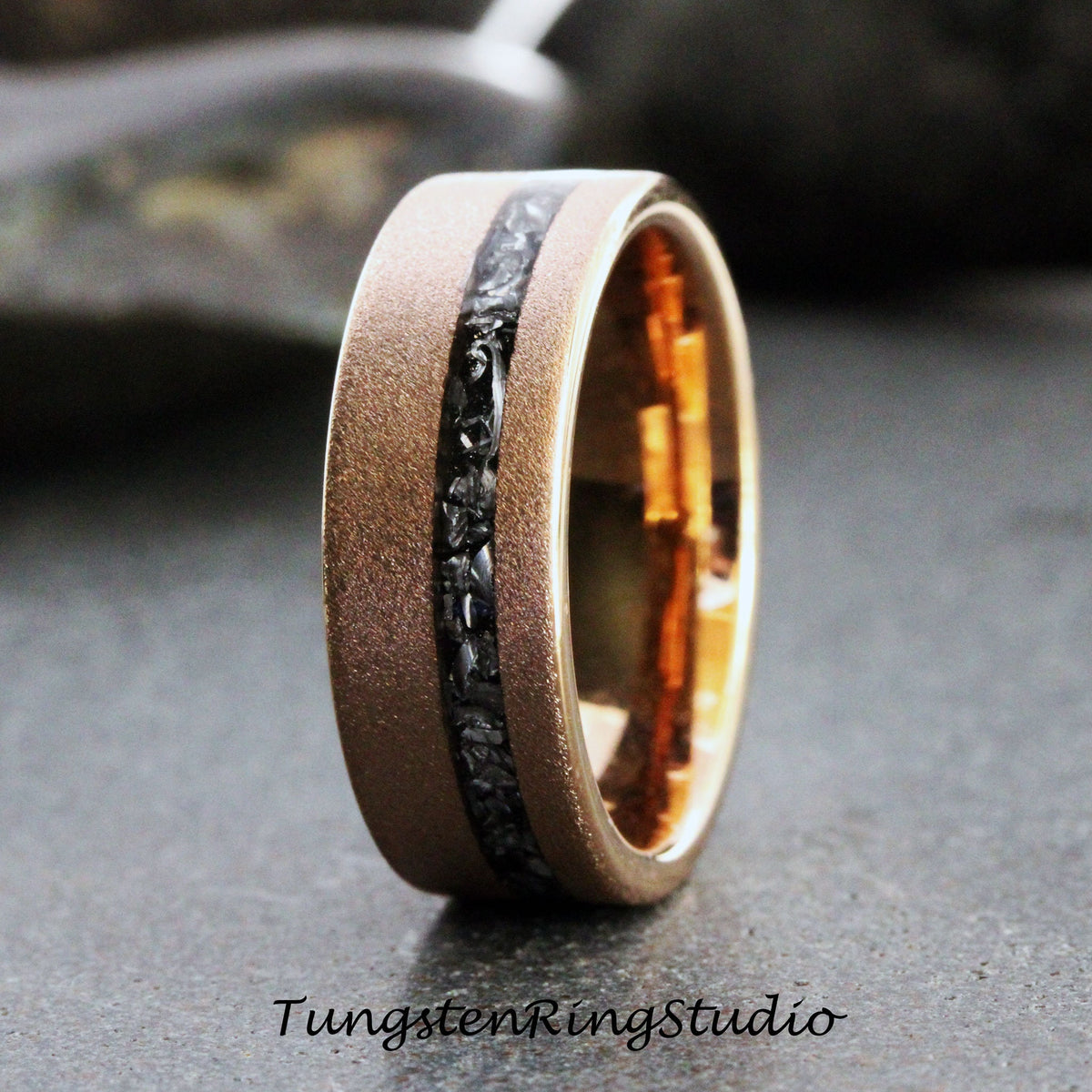 May include: A close-up of a gold-colored ring with a matte finish. The ring features a black, textured inlay running through the center. The text "TungstenRingstudio" is at the bottom of the image.