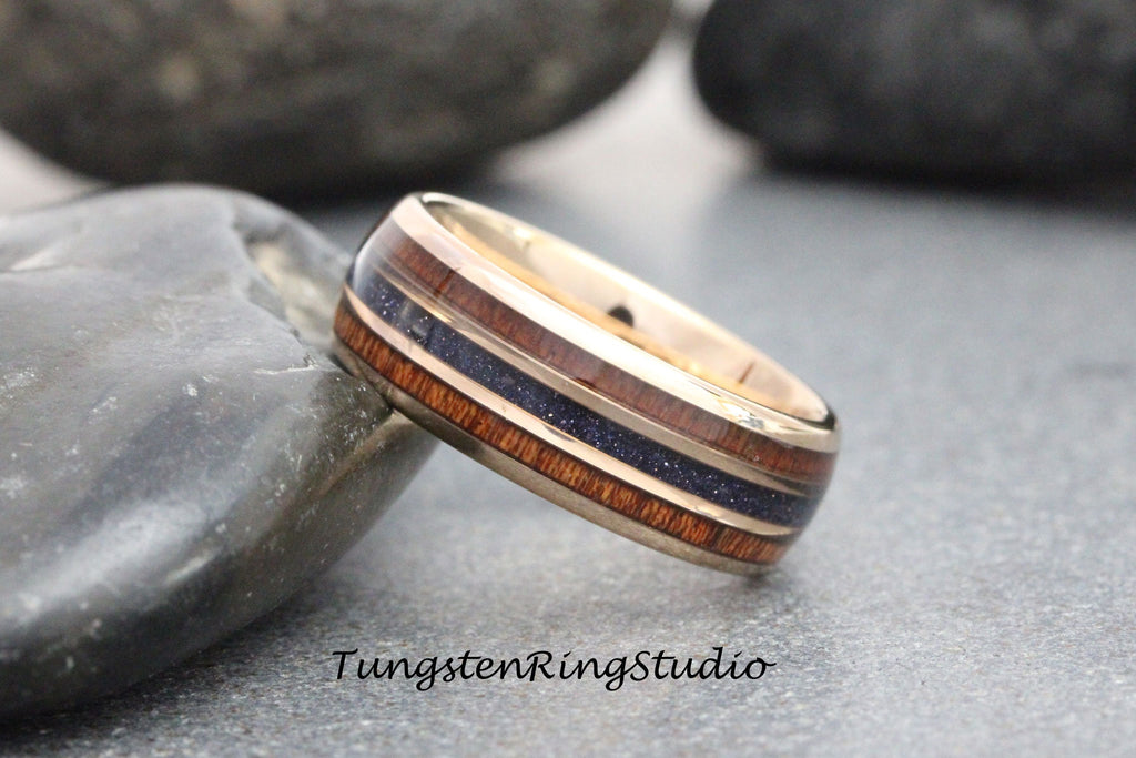May include: A rose gold tungsten carbide ring with a wooden inlay and blue glitter. The ring is on a gray stone background.