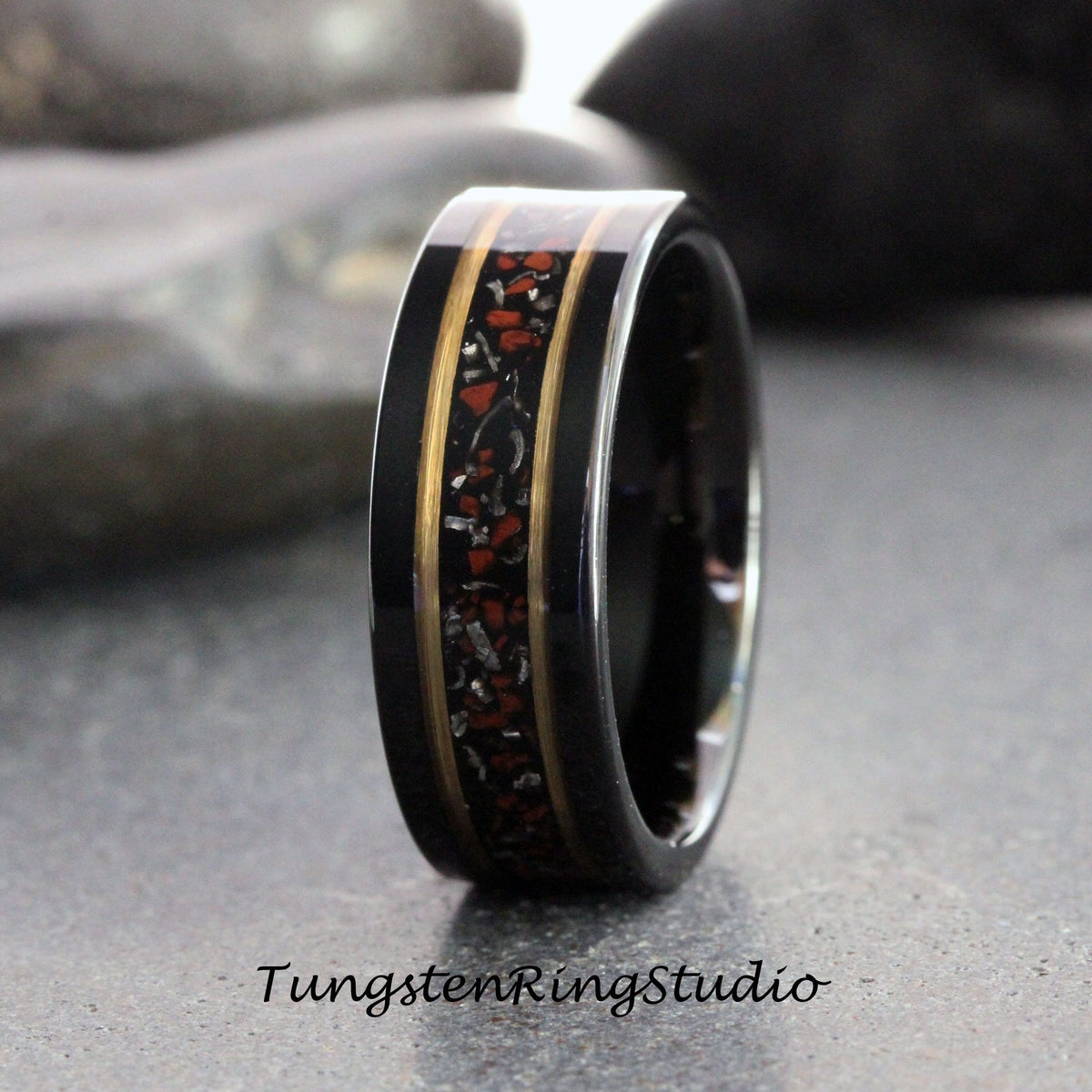 May include: A black tungsten carbide ring with a gold inlay and red and black speckled inlay. The ring is a wedding band.