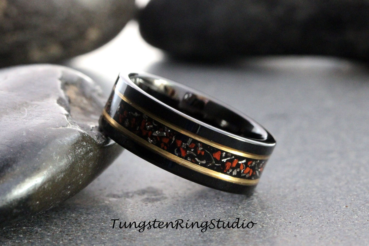 May include: Black tungsten carbide ring with a gold band and red and silver inlay. The ring is on a gray stone surface.