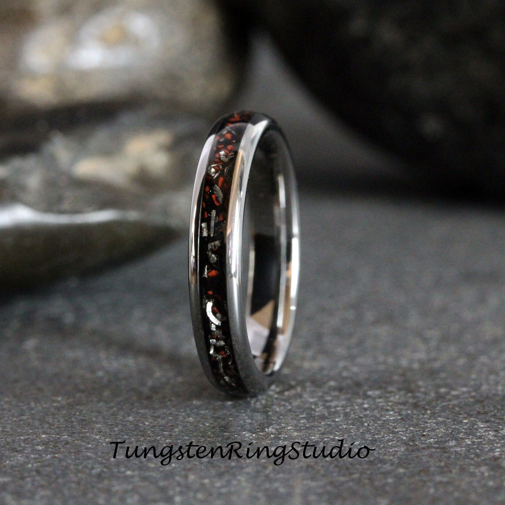 May include: A silver tungsten carbide ring with a black and red inlay. The inlay has a pattern of small, red and silver flecks.