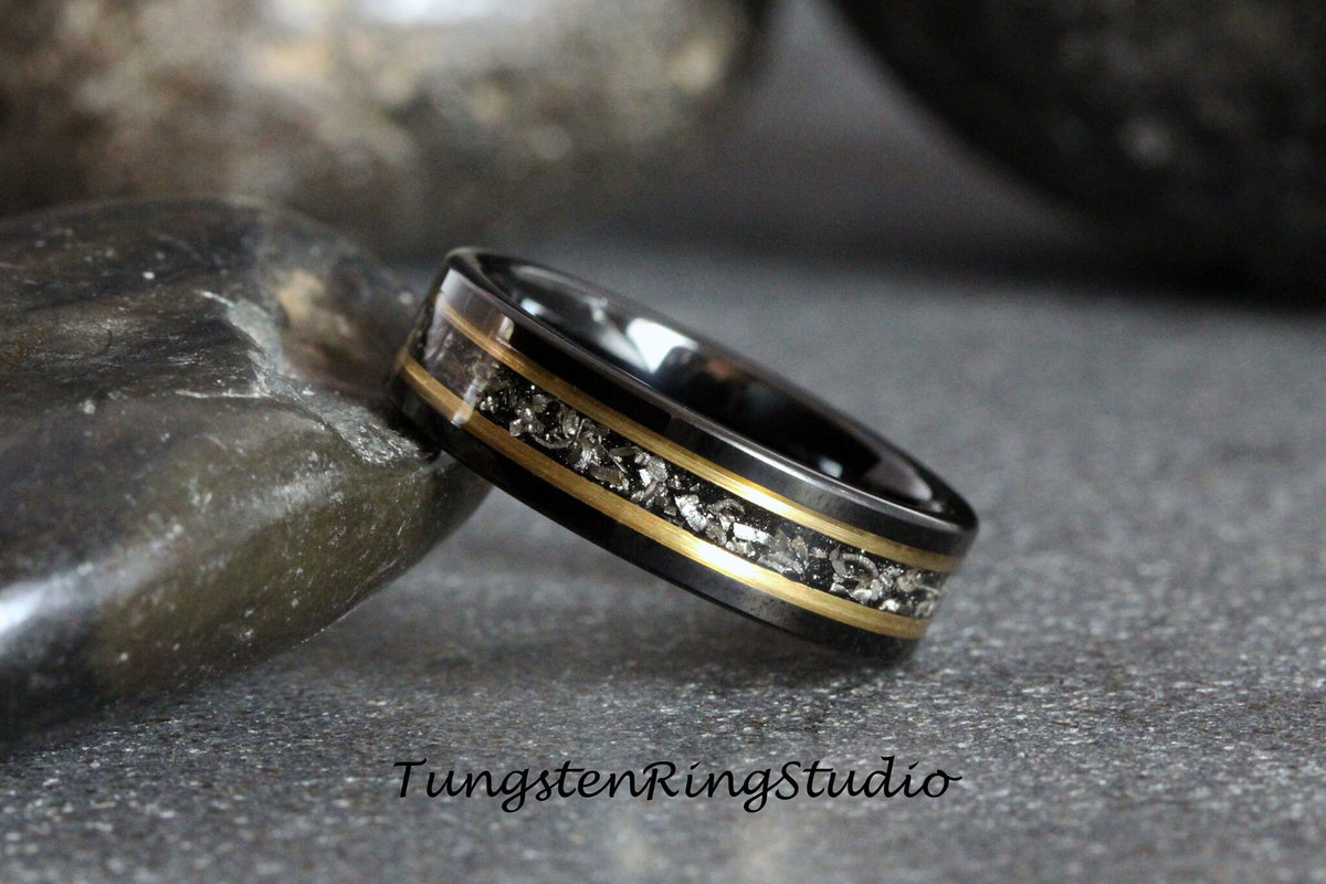 May include: A black tungsten ring with a gold inlay and a band of silver particles. The ring is on a gray surface.