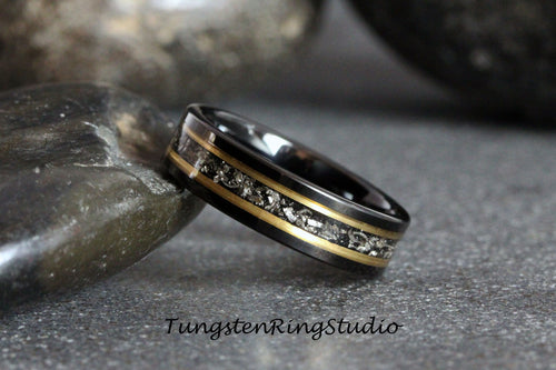 May include: A black tungsten ring with a gold inlay and a band of silver particles. The ring is on a gray surface.