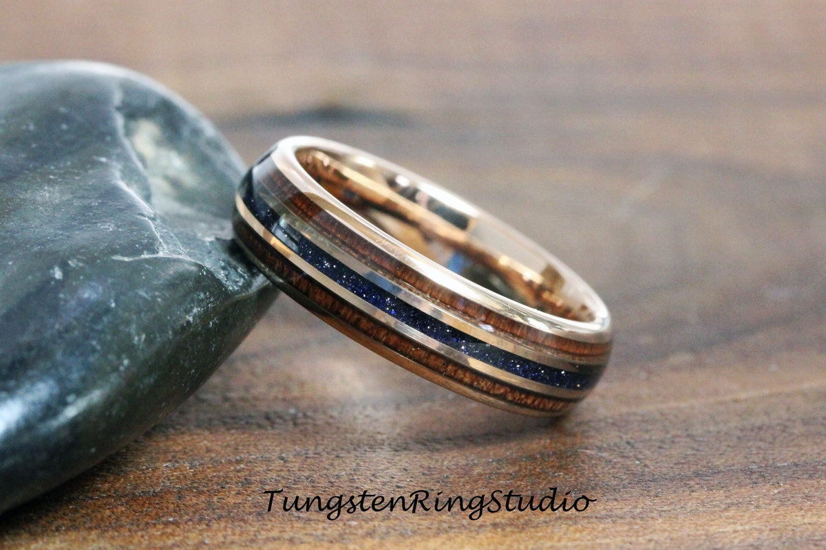 May include: A rose gold tungsten ring with a blue and brown wood inlay. The ring is on a gray rock and a brown wooden surface.