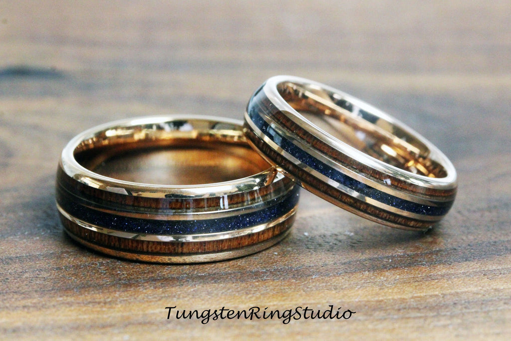 May include: Two gold tungsten carbide rings with a wooden inlay and blue glitter accents. The rings are stacked on top of each other.
