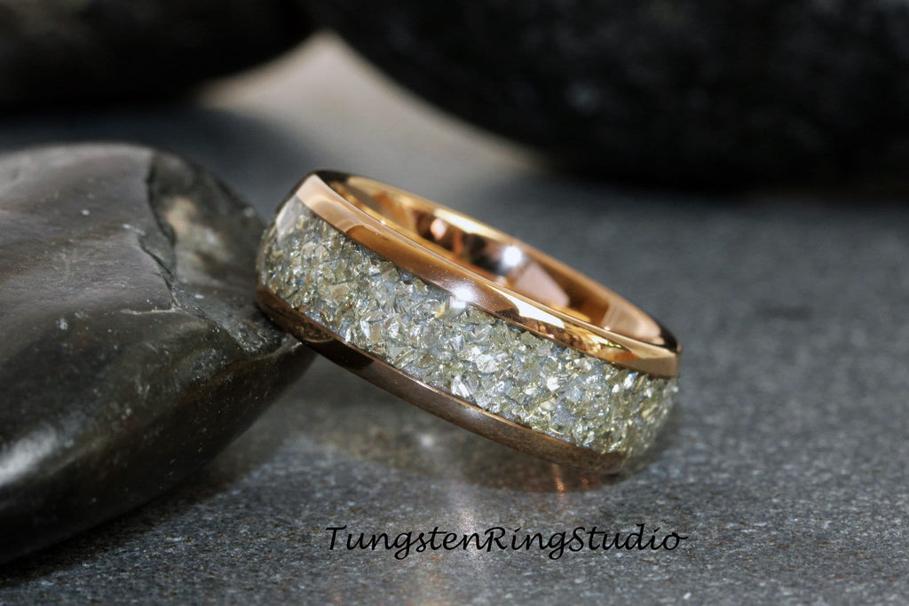 May include: A rose gold tungsten carbide ring with a band of embedded silver glitter. The ring is photographed on a gray background with two dark gray rocks.
