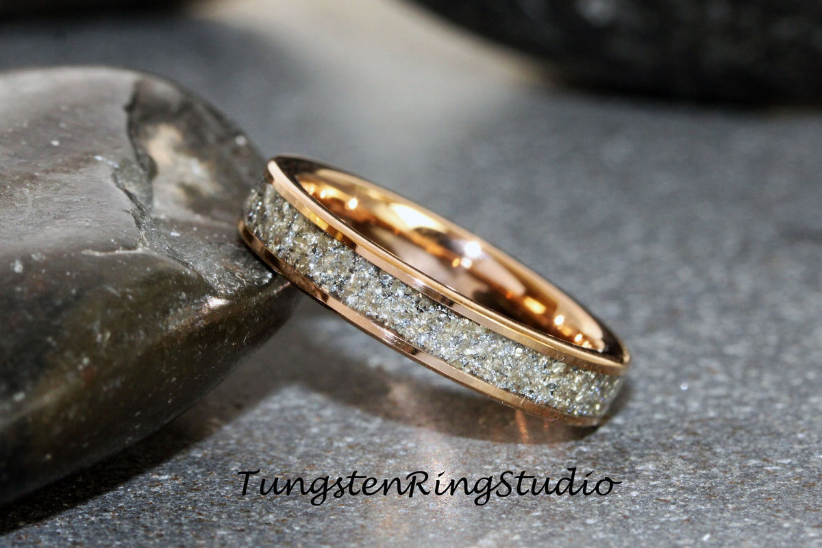 May include: A rose gold tungsten carbide ring with a silver inlay. The inlay is made of a textured material that resembles sand.