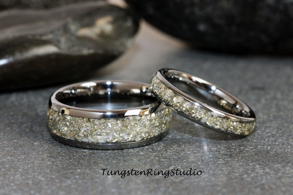 May include: Two silver tungsten carbide rings with a band of small, light-colored stones. The rings are on a gray surface. TungstenRingStudio.