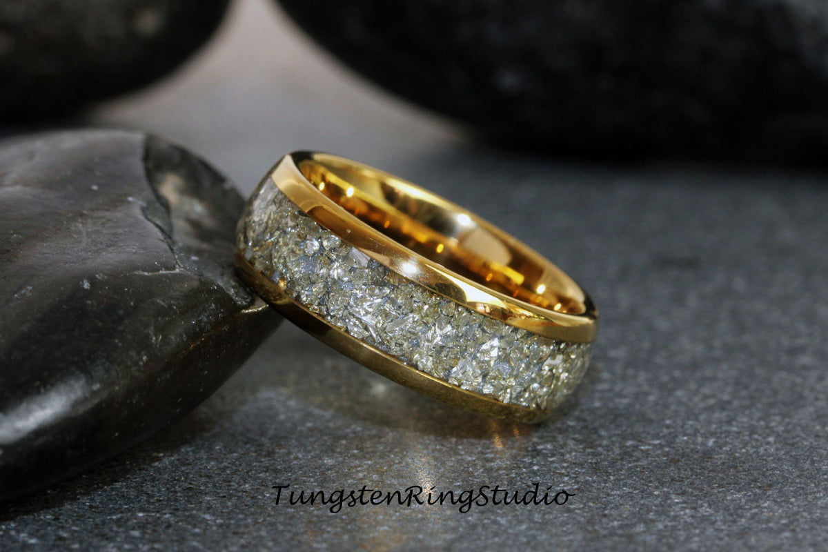 May include: A gold tungsten carbide ring with a white inlay of crushed meteorite. The ring is on a gray surface.