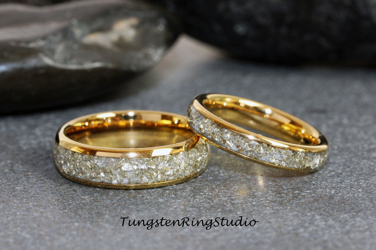 May include: Two gold tungsten carbide rings with a band of inlaid white diamonds. The rings are on a gray surface.