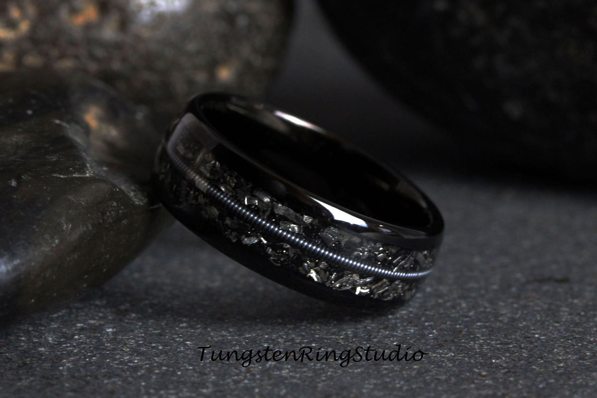 May include: A black tungsten carbide ring with a silver inlay and a black and silver speckled design. Tungsten Ring Studio is printed on the surface below the ring.