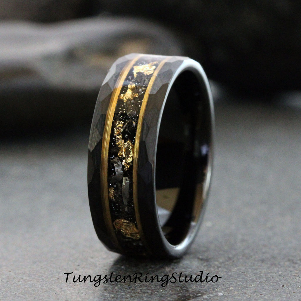 May include: A black tungsten carbide ring with a hammered finish and a gold inlay featuring black and gold flecks. Tungsten Ring Studio is printed below the ring.