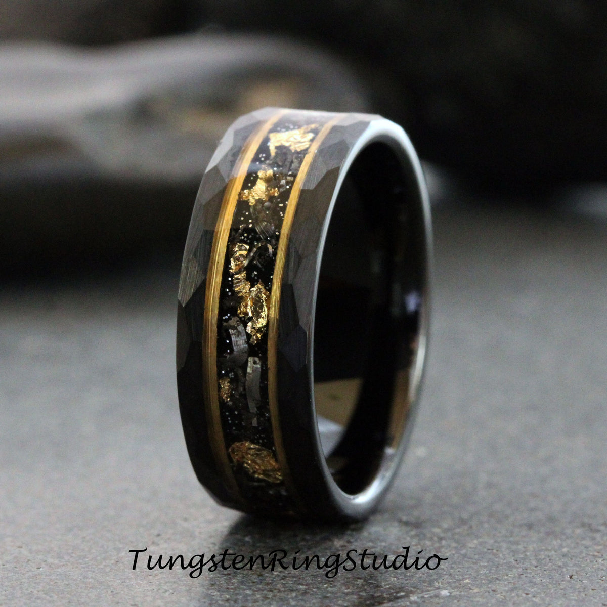 May include: A black tungsten carbide ring with a hammered finish and a gold inlay featuring black and gold flecks. Tungsten Ring Studio is printed below the ring.