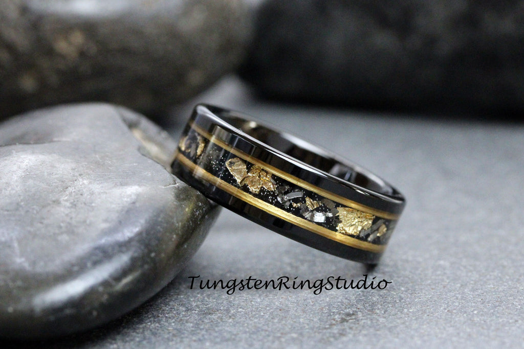 May include: Black tungsten carbide ring with a gold inlay and black and gold flecks. The ring is on a gray stone background. TungstenRingStudio.