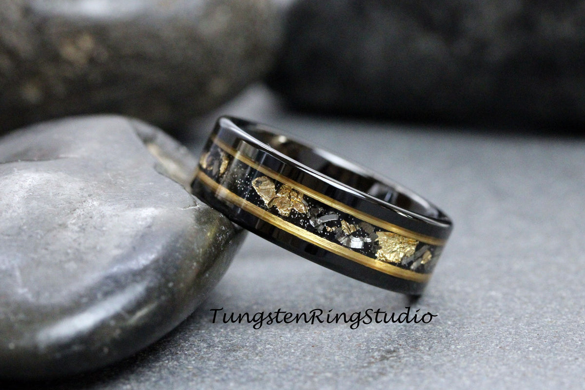 May include: Black tungsten carbide ring with a gold inlay and black and gold flecks. The ring is on a gray stone background. TungstenRingStudio.