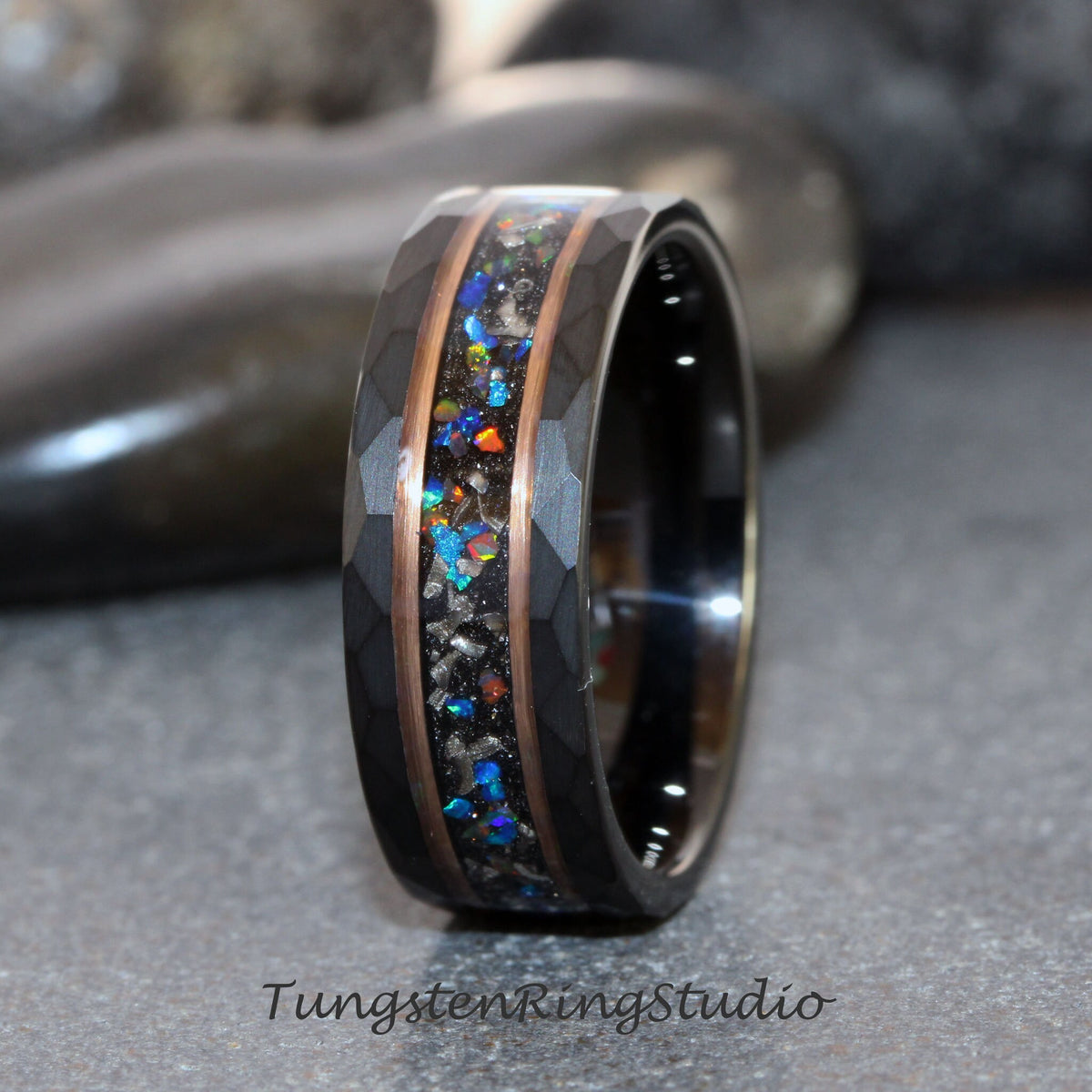 May include: A black tungsten ring with a hammered finish. The ring features a central inlay of colorful, iridescent material, framed by copper-colored bands. The text "TungstenRingStudio" is at the bottom.