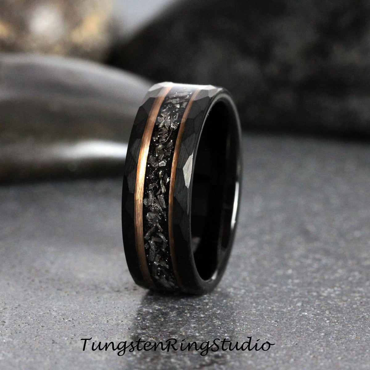 May include: A black tungsten carbide ring with a hammered finish and a rose gold inlay. The inlay features a pattern of small, silver-colored particles.