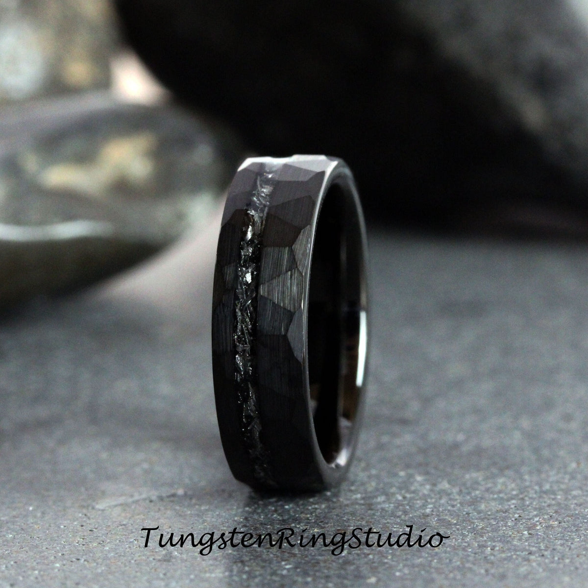 May include: A black tungsten carbide ring with a hammered finish and a thin line of silver inlay. The ring is shown on a gray background.