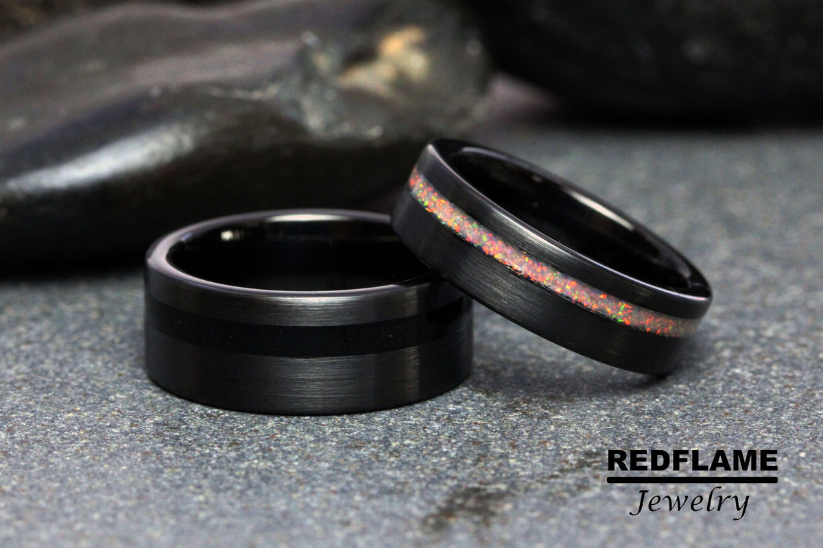 May include: Two black tungsten carbide rings. One ring has a thin black inlay, the other has a thin inlay of iridescent opal.
