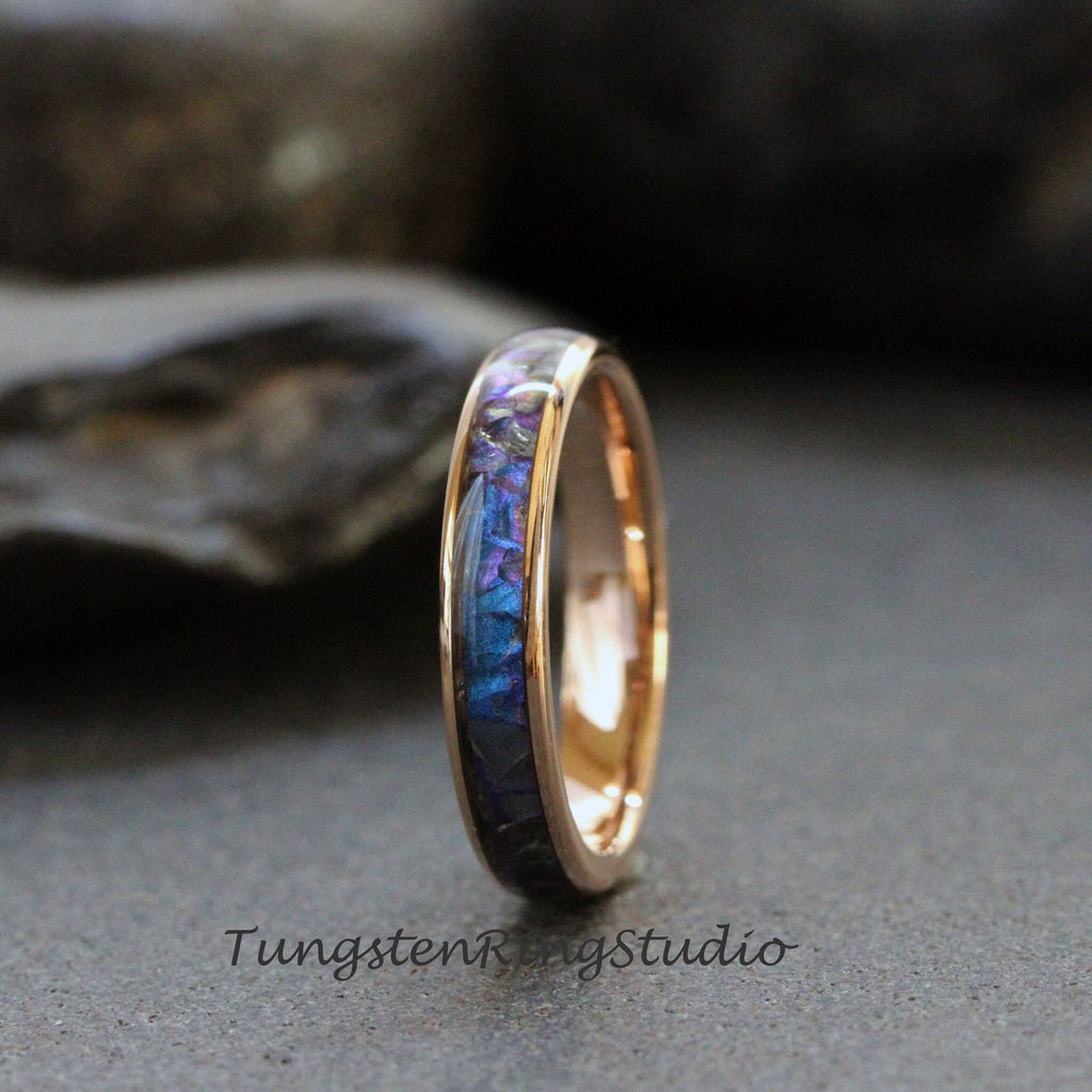 May include: A rose gold tungsten carbide ring with a blue and purple inlay. The inlay is made of crushed meteorite and is set in a polished ring band.
