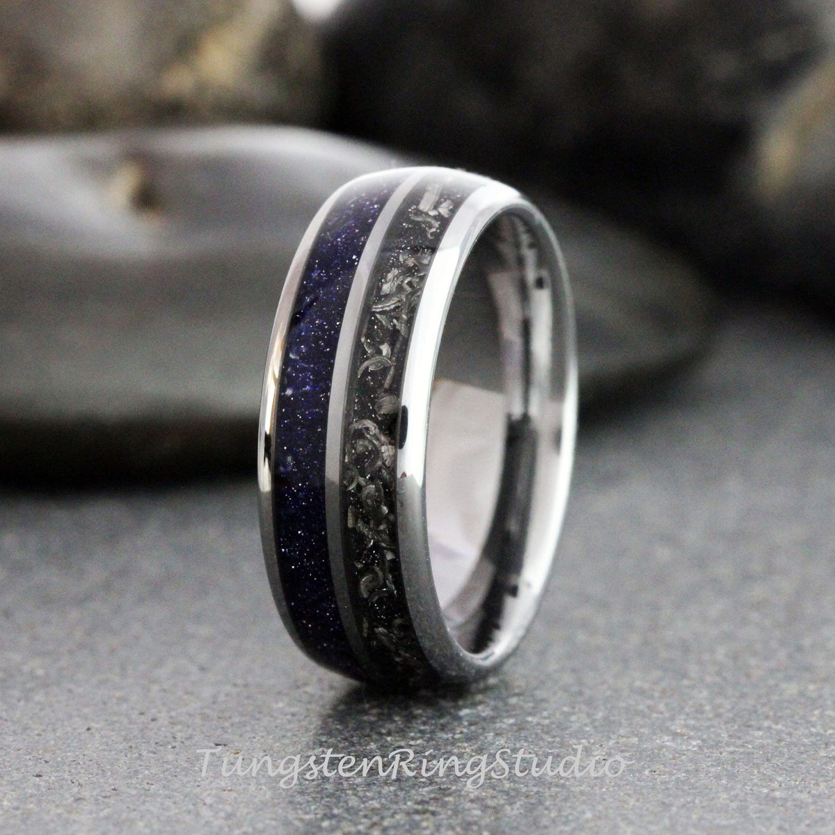 May include: A polished silver ring with a dark blue, speckled inlay and a dark gray, textured inlay. The ring has a shiny, metallic finish and is set against a blurred background. The text "TungstenRingStudio" is visible.