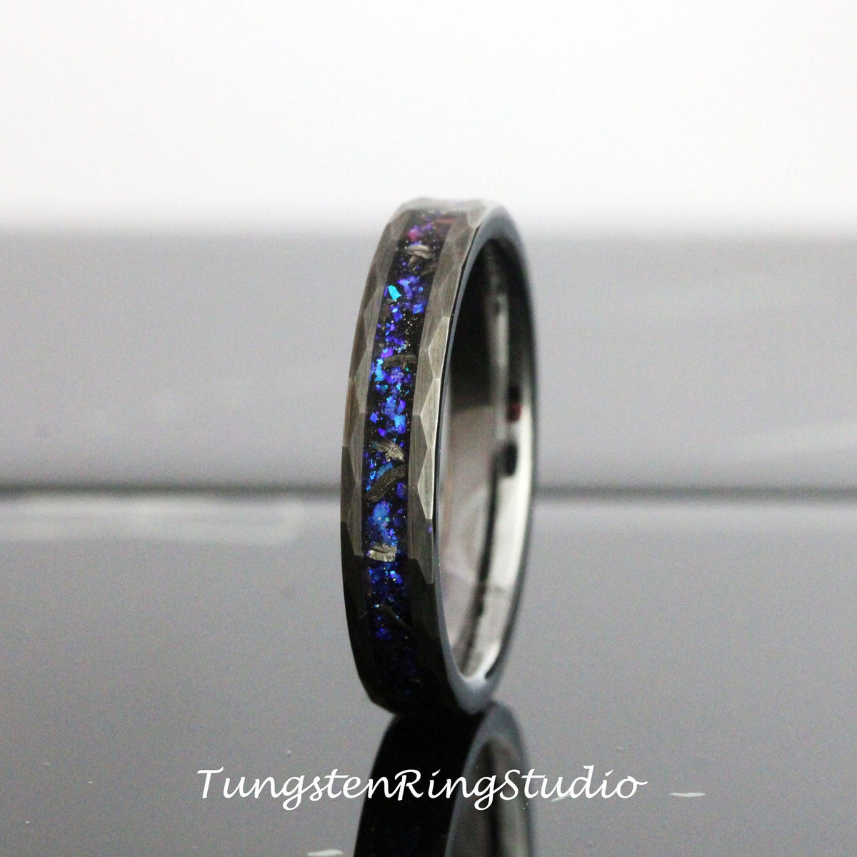 May include: A black tungsten carbide ring with a blue and purple iridescent inlay. The ring has a hammered finish and a beveled edge.