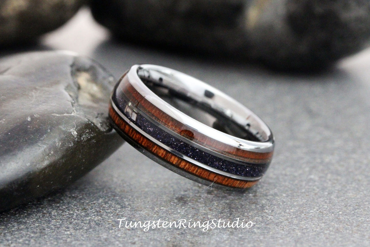 May include: A silver tungsten ring with a dark purple and brown wood inlay. The ring is on a gray rock.