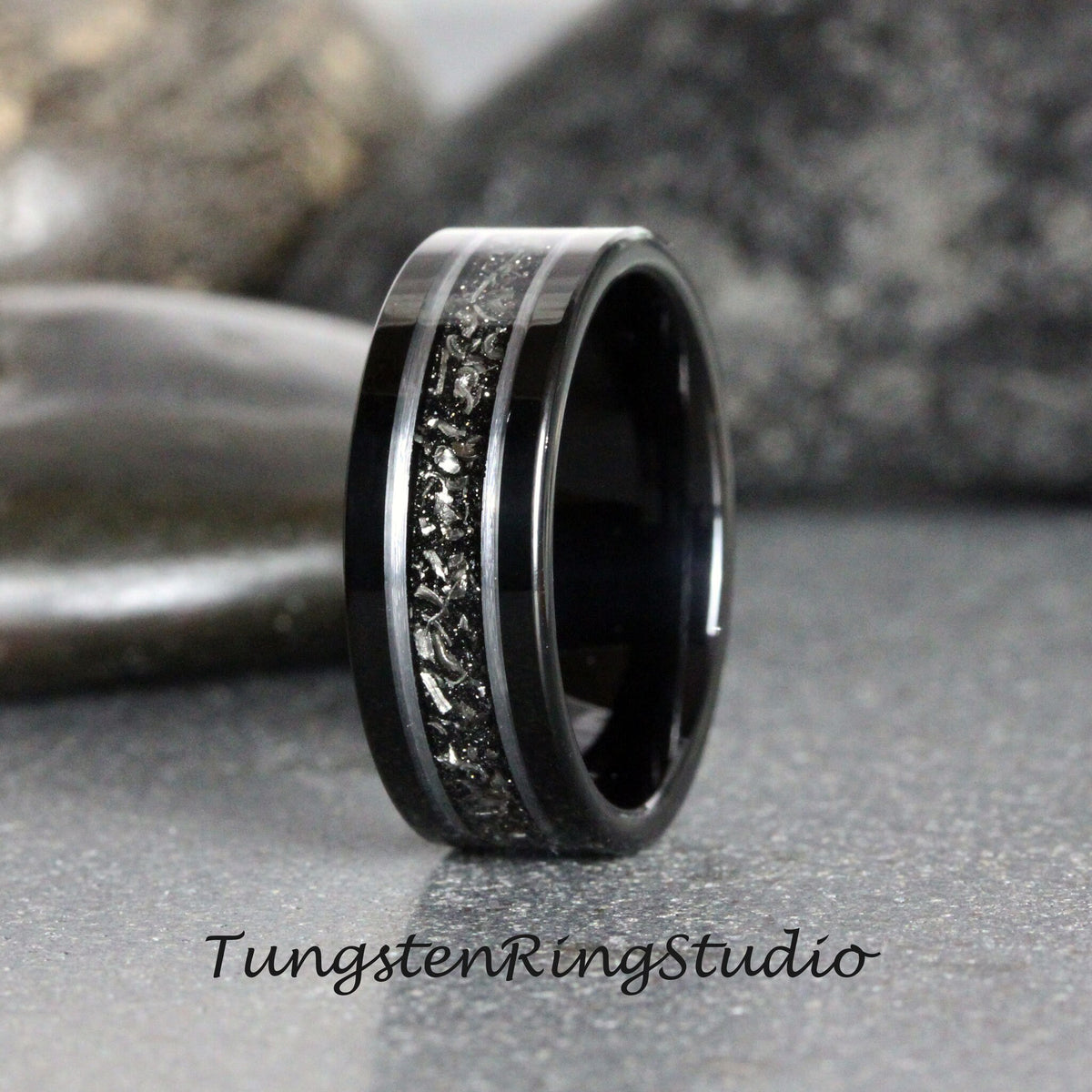 May include: A black tungsten carbide ring with a silver inlay and a black and silver speckled design. Tungsten Ring Studio is printed on the surface below the ring.